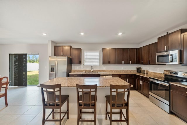 kitchen featuring a kitchen bar, appliances with stainless steel finishes, light stone countertops, dark brown cabinetry, and a kitchen island