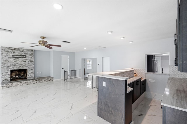 kitchen with a stone fireplace, ceiling fan, kitchen peninsula, and light stone counters