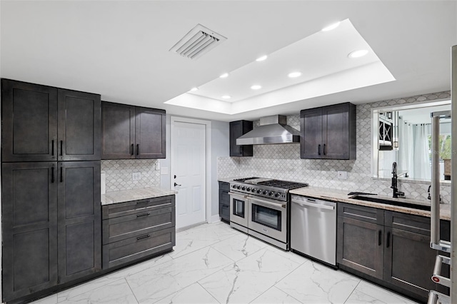 kitchen featuring backsplash, stainless steel appliances, a tray ceiling, sink, and wall chimney range hood