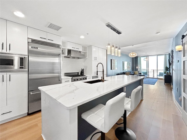 kitchen featuring a center island with sink, sink, built in appliances, decorative light fixtures, and white cabinetry