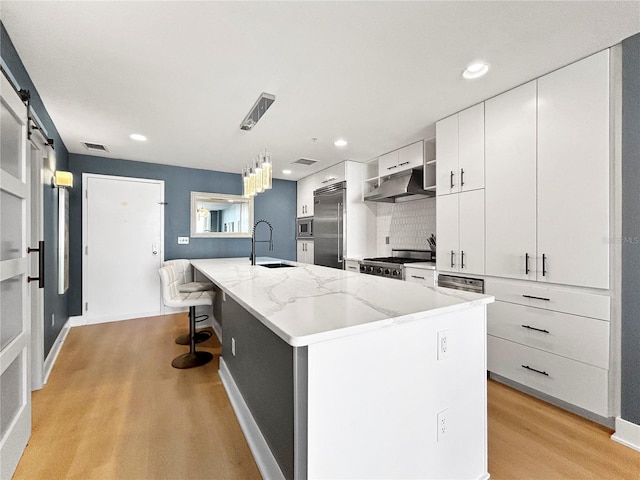 kitchen with a kitchen island with sink, sink, built in appliances, a barn door, and white cabinetry