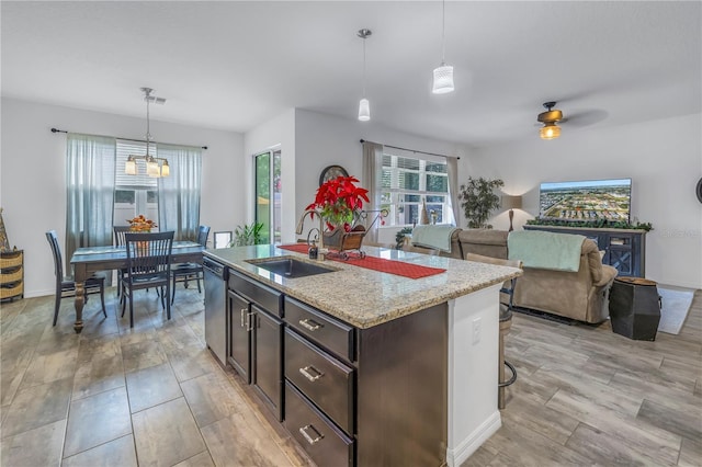 kitchen with light stone counters, ceiling fan, a kitchen island with sink, dishwasher, and hanging light fixtures