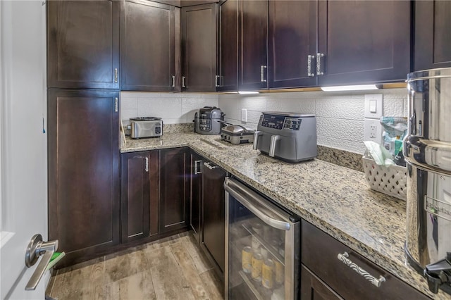 kitchen with light stone countertops, wine cooler, decorative backsplash, dark brown cabinets, and light wood-type flooring