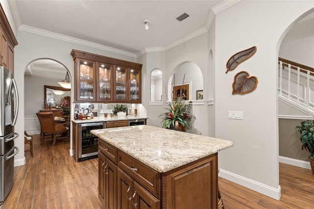 bar with wine cooler, wood-type flooring, stainless steel refrigerator, ornamental molding, and light stone counters