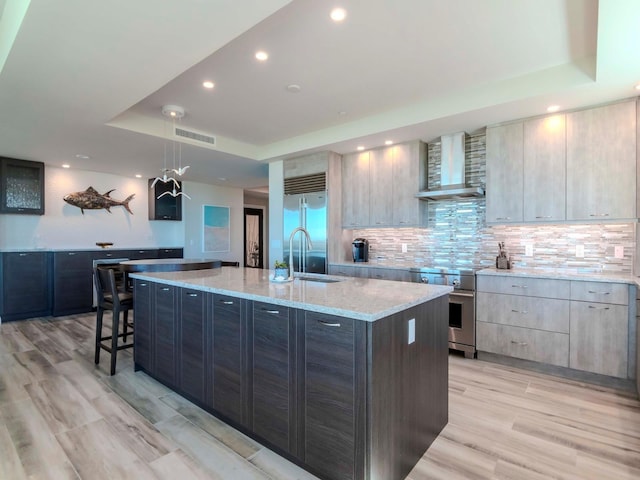 kitchen featuring pendant lighting, wall chimney exhaust hood, an island with sink, light stone counters, and stainless steel appliances