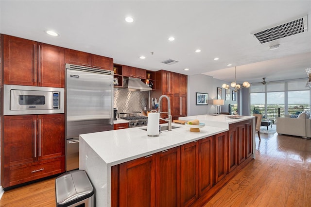 kitchen with sink, backsplash, built in appliances, a kitchen island with sink, and light wood-type flooring