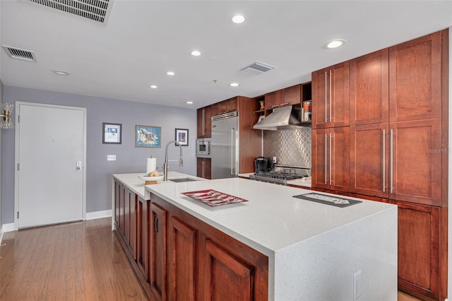 kitchen with sink, built in appliances, decorative backsplash, light wood-type flooring, and an island with sink