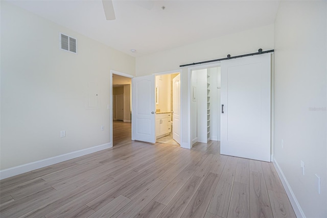 unfurnished bedroom with ceiling fan, a barn door, light wood-type flooring, and ensuite bath