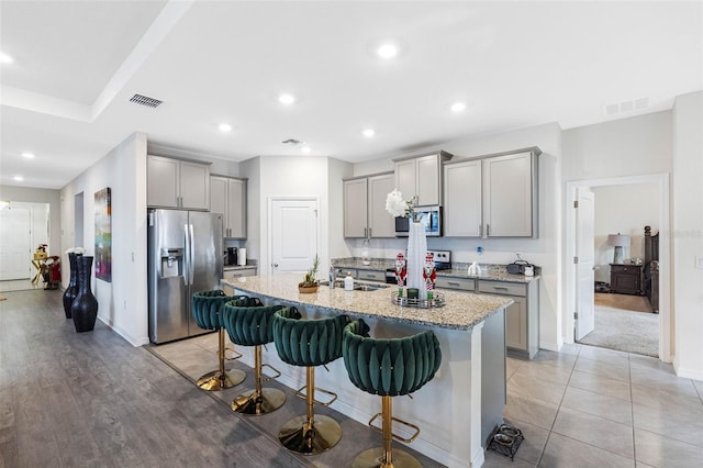 kitchen featuring a kitchen breakfast bar, light stone counters, stainless steel appliances, a kitchen island with sink, and gray cabinets