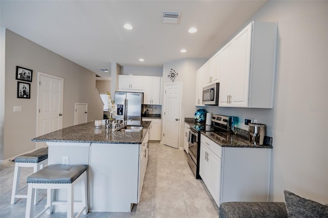 kitchen with white cabinets, dark stone counters, stainless steel appliances, and a center island with sink