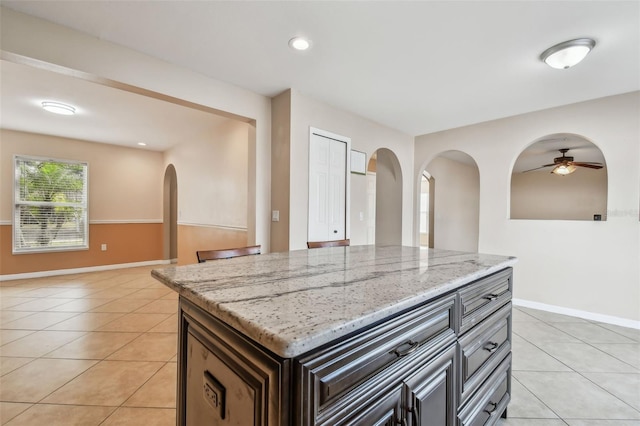 kitchen featuring ceiling fan, a kitchen island, light stone counters, dark brown cabinets, and light tile patterned flooring