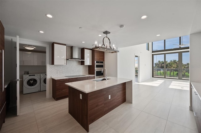 kitchen with a kitchen island with sink, black electric stovetop, wall chimney exhaust hood, washer and dryer, and decorative light fixtures