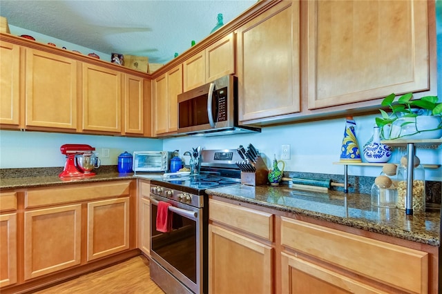 kitchen with light wood-type flooring, a textured ceiling, appliances with stainless steel finishes, and dark stone counters