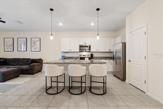 kitchen with dark stone counters, stainless steel appliances, a kitchen island with sink, decorative light fixtures, and white cabinetry