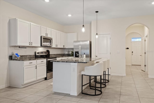 kitchen featuring light tile patterned floors, dark stone countertops, a center island with sink, white cabinets, and appliances with stainless steel finishes