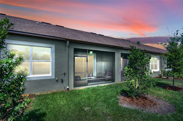 back of property at dusk with a sunroom, a shingled roof, a lawn, and stucco siding