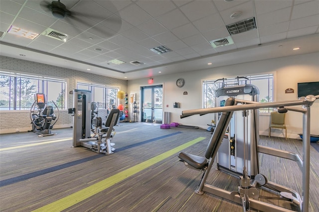 exercise room featuring ceiling fan, carpet floors, a drop ceiling, and brick wall