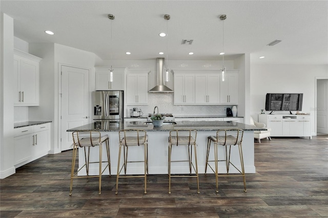 kitchen with white cabinetry, wall chimney exhaust hood, stainless steel fridge, an island with sink, and pendant lighting