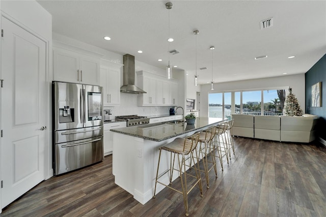 kitchen with wall chimney exhaust hood, an island with sink, dark stone counters, white cabinets, and appliances with stainless steel finishes