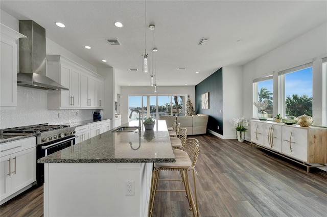 kitchen featuring wall chimney exhaust hood, a kitchen island with sink, sink, stainless steel stove, and white cabinetry