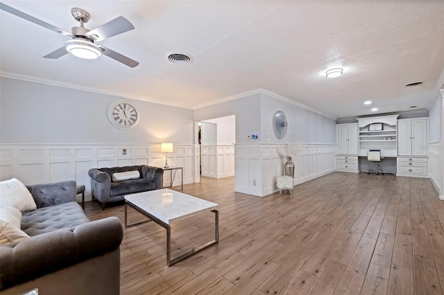 living room with built in desk, light wood-type flooring, ornamental molding, ceiling fan, and a textured ceiling