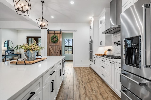 kitchen with white cabinetry, stainless steel appliances, wall chimney range hood, a barn door, and decorative light fixtures