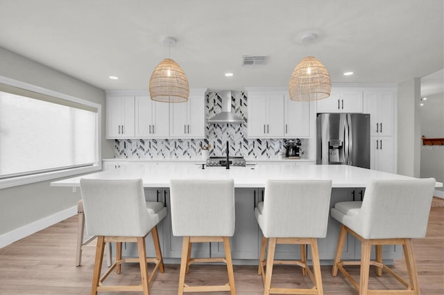 kitchen with appliances with stainless steel finishes, wall chimney range hood, a large island with sink, decorative light fixtures, and white cabinets