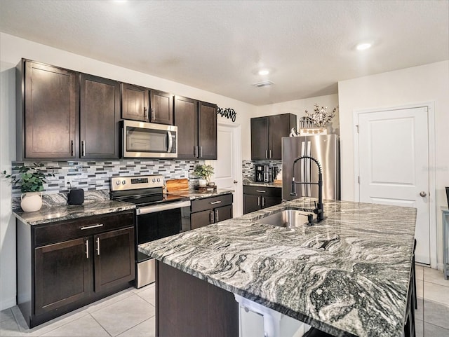 kitchen with stone counters, sink, a kitchen island with sink, stainless steel appliances, and dark brown cabinets