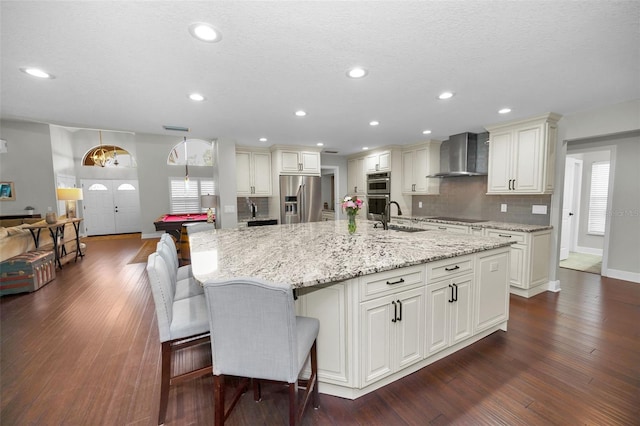 kitchen with tasteful backsplash, a large island with sink, stainless steel appliances, light stone countertops, and wall chimney exhaust hood