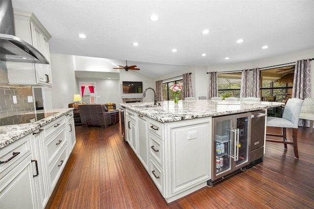 kitchen featuring wine cooler, black electric cooktop, a kitchen island with sink, decorative backsplash, and wall chimney range hood