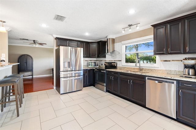 kitchen with sink, wall chimney exhaust hood, ceiling fan, light stone counters, and stainless steel appliances