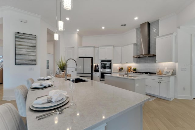 kitchen with a center island with sink, wall chimney range hood, a sink, stainless steel double oven, and ornamental molding