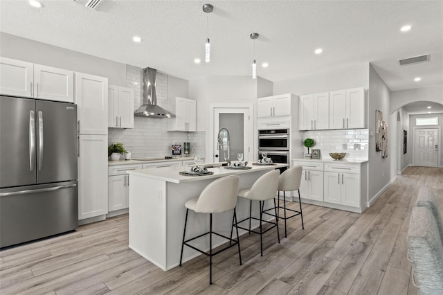 kitchen with white cabinets, backsplash, a kitchen island with sink, stainless steel appliances, and wall chimney exhaust hood