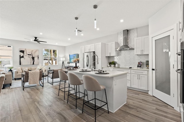 kitchen featuring stainless steel refrigerator, wall chimney exhaust hood, a center island with sink, and white cabinets