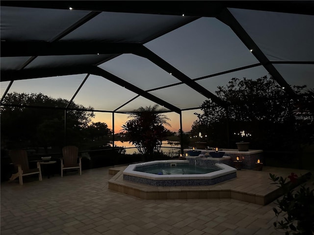 pool at dusk featuring an in ground hot tub, a patio area, glass enclosure, and a water view