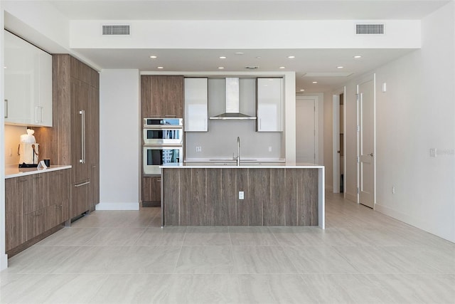 kitchen with wall chimney exhaust hood, stainless steel double oven, sink, light tile patterned floors, and a center island with sink