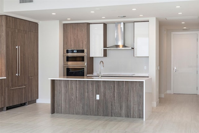 kitchen featuring wall chimney exhaust hood, stainless steel double oven, sink, light tile patterned floors, and a center island with sink