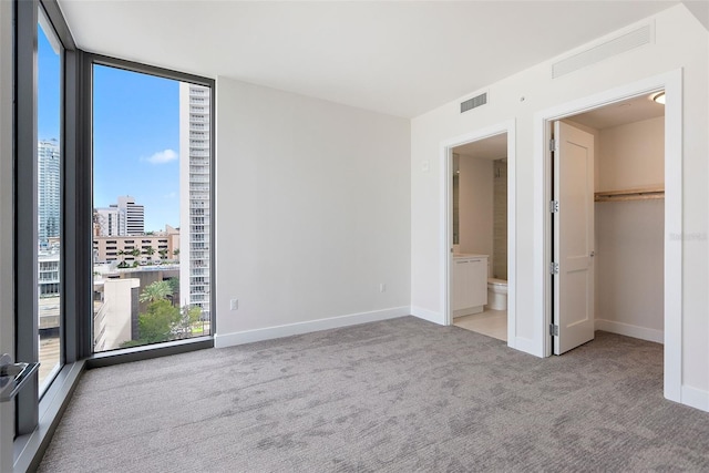 unfurnished bedroom featuring light carpet, ensuite bath, a spacious closet, a wall of windows, and a closet