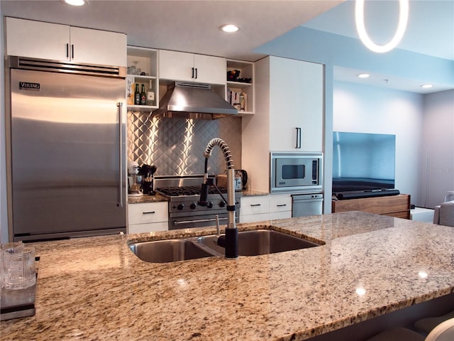 kitchen with wall chimney range hood, built in appliances, decorative backsplash, light stone counters, and white cabinetry