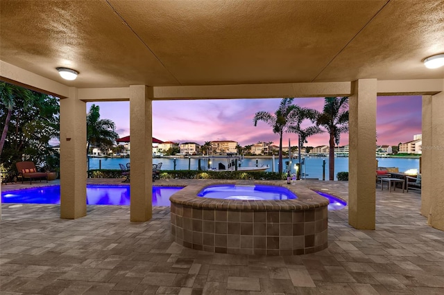 pool at dusk featuring an in ground hot tub, a water view, and a patio