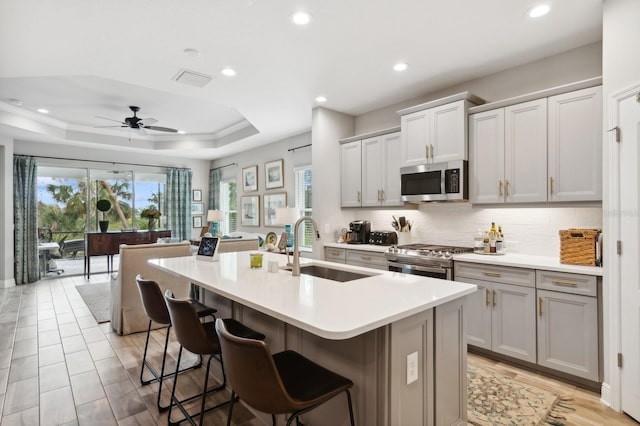 kitchen with a raised ceiling, sink, a kitchen island with sink, appliances with stainless steel finishes, and a kitchen breakfast bar