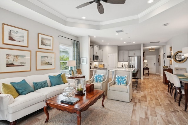 living room featuring light hardwood / wood-style floors, sink, ornamental molding, ceiling fan, and a tray ceiling