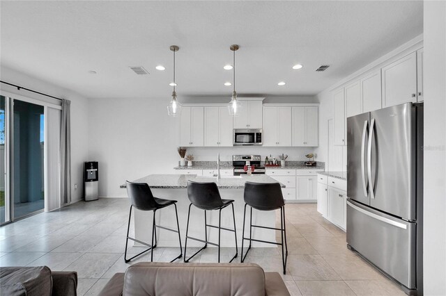 kitchen with white cabinets, hanging light fixtures, an island with sink, light stone counters, and stainless steel appliances