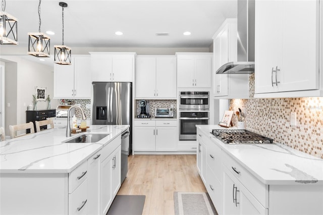 kitchen featuring appliances with stainless steel finishes, wall chimney exhaust hood, decorative light fixtures, white cabinetry, and an island with sink