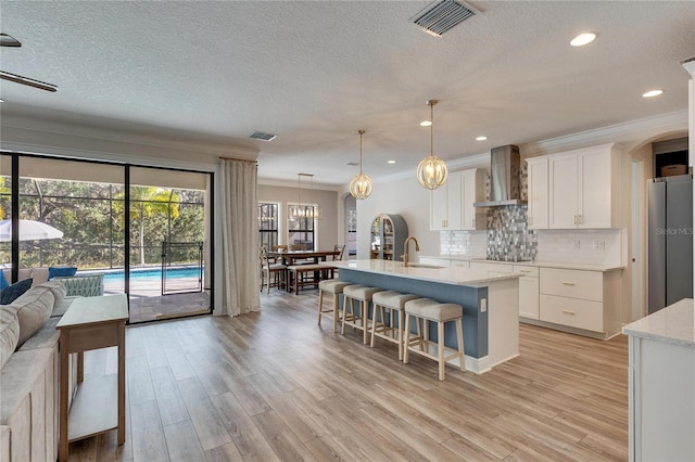 kitchen with wall chimney exhaust hood, ornamental molding, an island with sink, decorative light fixtures, and white cabinetry