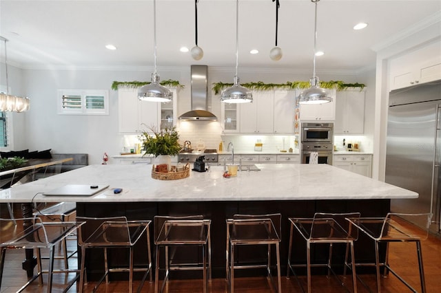 kitchen with appliances with stainless steel finishes, decorative light fixtures, a large island with sink, light stone counters, and wall chimney range hood