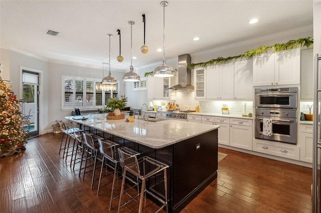 kitchen featuring a spacious island, wall chimney exhaust hood, appliances with stainless steel finishes, a kitchen breakfast bar, and light stone countertops