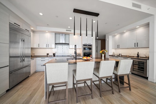 kitchen with stainless steel appliances, wall chimney range hood, white cabinets, wine cooler, and hanging light fixtures