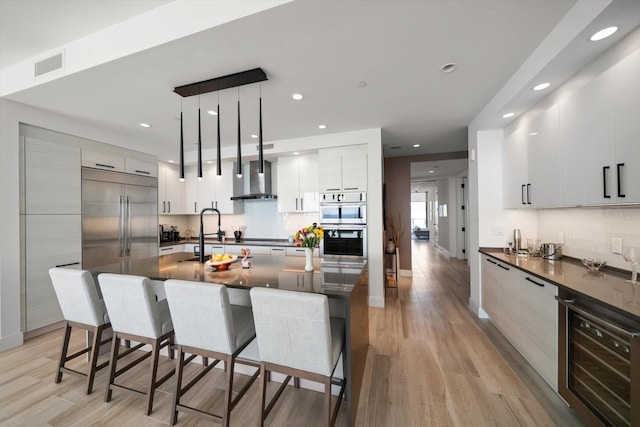 kitchen featuring white cabinetry, wall chimney exhaust hood, beverage cooler, hanging light fixtures, and a kitchen breakfast bar