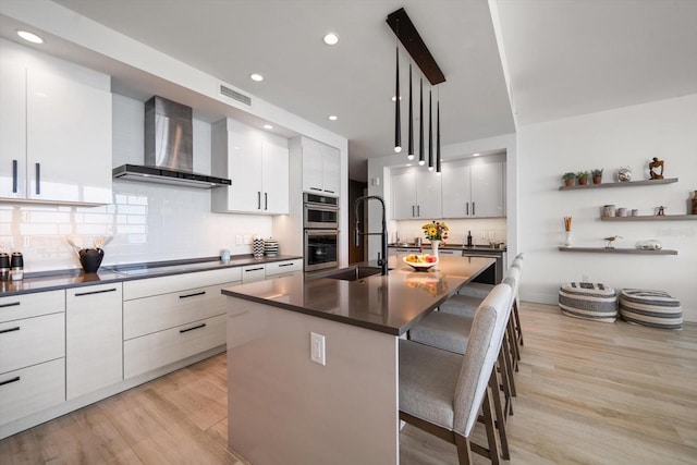 kitchen featuring a breakfast bar, wall chimney range hood, hanging light fixtures, an island with sink, and stovetop